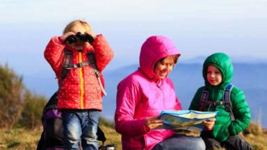 Une mère avec sa fille et son fils à la montagne, habillés avec des vêtements chauds, la petite fille regarde dans une paire de jumelles et la mère regarde une carte.